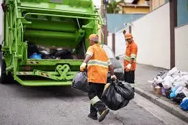Two sanitation workers in orange uniforms collecting black trash bags near a green garbage truck.